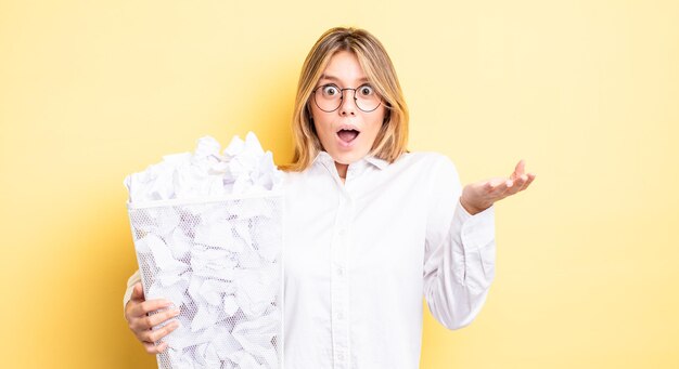 Bonita chica rubia asombrada, conmocionada y atónita con una sorpresa increíble. concepto de basura de bolas de papel