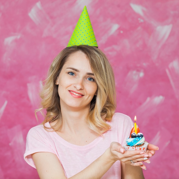 Bonita chica rubia en gorro de cumpleaños mantenga cupcake con vela única