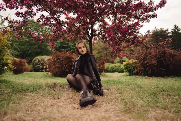 Bonita chica rubia de negro se sienta en el suelo en el parque y se relaja bajo un árbol de follaje rosa