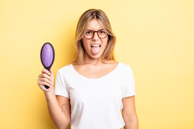 Bonita chica rubia que se siente disgustada e irritada y con la lengua fuera. concepto de cepillo de pelo