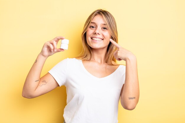 Bonita chica rubia sonriendo con confianza apuntando a su propia amplia sonrisa. concepto de botella de pastillas