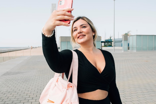 Bonita chica rubia de talla grande tomando una selfie con su teléfono celular frente a la playa vestida con ropa deportiva