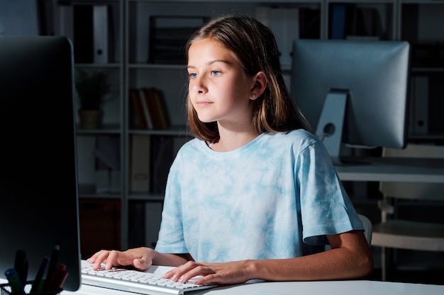 Bonita colegiala en camiseta mirando la pantalla del ordenador mientras la creación de redes en el aula oscura después de las lecciones
