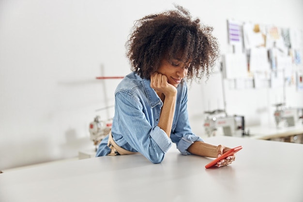 Bonita diseñadora afroamericana en camisa de mezclilla utiliza teléfono móvil en un espacioso taller de costura