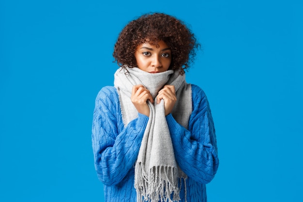 Bonita e sombria jovem adorável afro-americana com corte de cabelo afro, enrole-se com um lenço quente, sentindo frio ou frio no inverno, em pé na parede azul, esperando alguém consertar o aquecedor