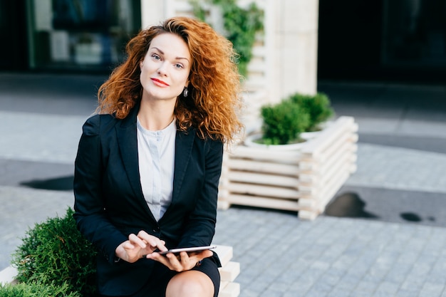 Foto bonita empresária com cabelos cacheados, pele pura e lábios vermelhos, vestindo traje formal enquanto está sentado no café ao ar livre