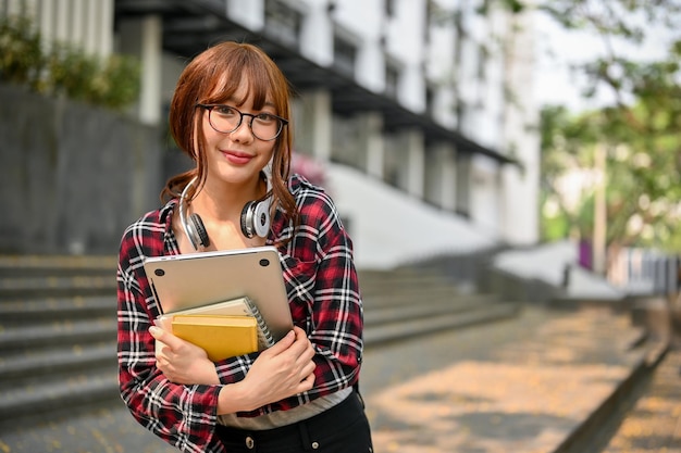 Bonita estudiante asiática sosteniendo su laptop y su libro escolar parados afuera del edificio