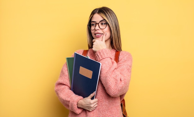 Bonita estudiante mujer sonriendo con una expresión feliz y segura con la mano en la barbilla. libros y mochila