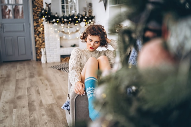 Bonita y feliz morena sentada en suéter caliente en la silla con luces de Navidad detrás