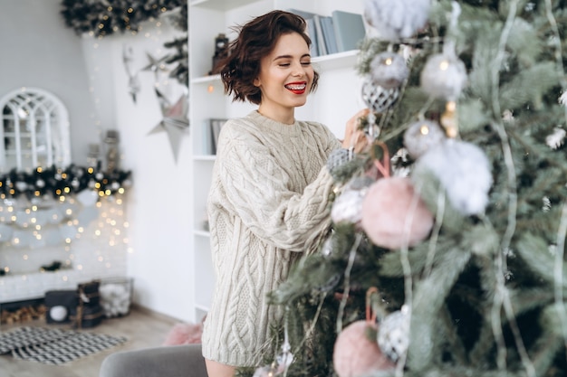 Bonita y feliz morena en suéter cálido decorar el árbol de Navidad en casa