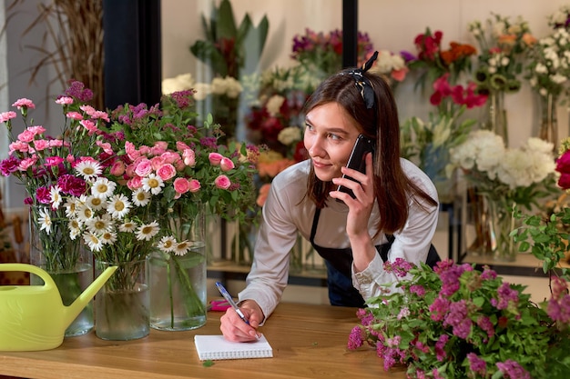 Bonita floristería aceptando pedidos por teléfono y tomando notas en el cuaderno en la florería. Asistente de mujer o propietario en estudio de diseño floral, haciendo decoraciones. Servicio de entrega de flores.