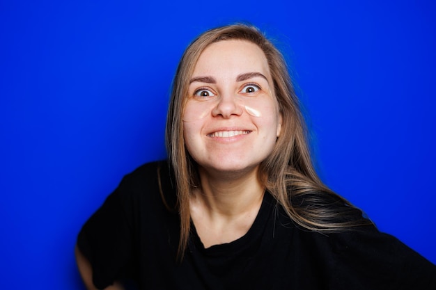 Bonita jovem loira com pele lisa natural sem maquiagem posando feliz e sorrindo para a câmera em pé na camiseta sobre fundo azul Conceito de beleza tapa-olhos para mulheres