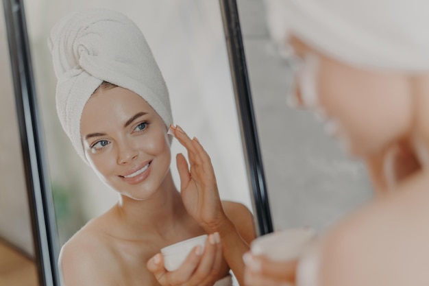 Bonita jovem sorridente aplica creme de beleza no rosto sorri agradavelmente tem maquiagem mínima usa toalha enrolada na cabeça olha em poses de espelho no banheiro Conceito de tratamentos de beleza de bem-estar