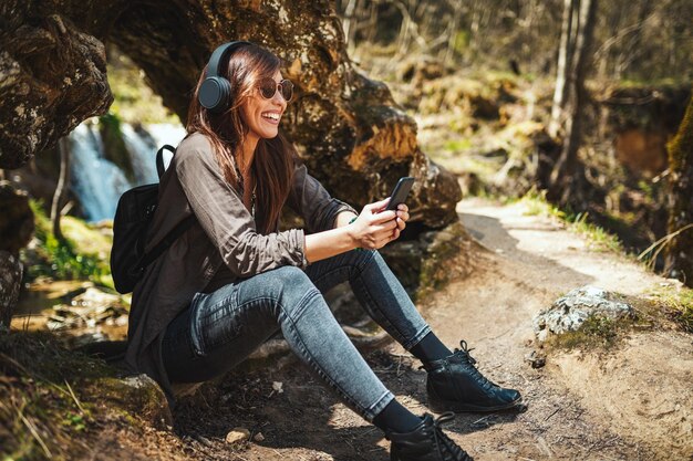 Bonita jovem sorridente, com smartphone na mão e fones de ouvido na cabeça, ouvindo música e apreciando a paisagem nas montanhas, olhando para a bela cachoeira.
