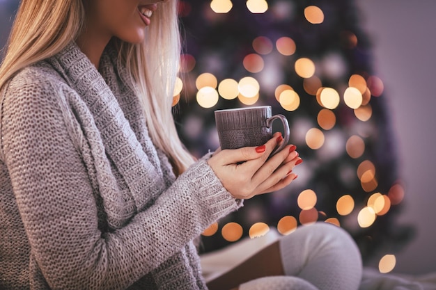 Bonita jovem sorridente sentada na cama e segurando uma xícara de café ou chá, rodeada de luzes de Natal.