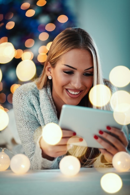 Bonita jovem sorridente usando tablet e feliz sorrindo durante as férias de Natal aconchegantes em casa.