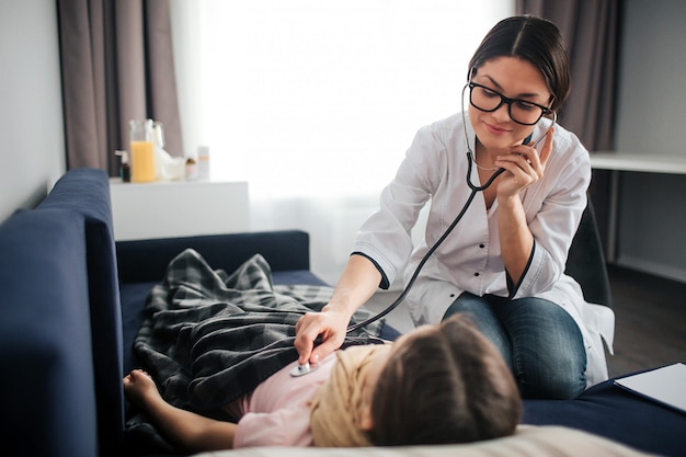 Bonita joven doctora escucha la respiración del niño a través del estetoscopio. Ella sonríe y mira a la chica. El doctor se sienta además. Niño recostado en el sofá.