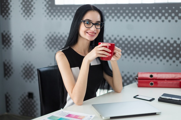 Bonita, joven empresaria en vestido negro y anteojos se sienta a la mesa y sostiene la taza roja