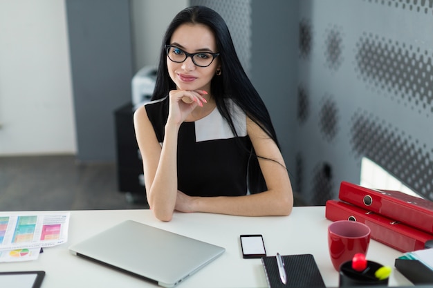 Bonita, joven empresaria en vestido negro y anteojos se sienta a la mesa y trabaja