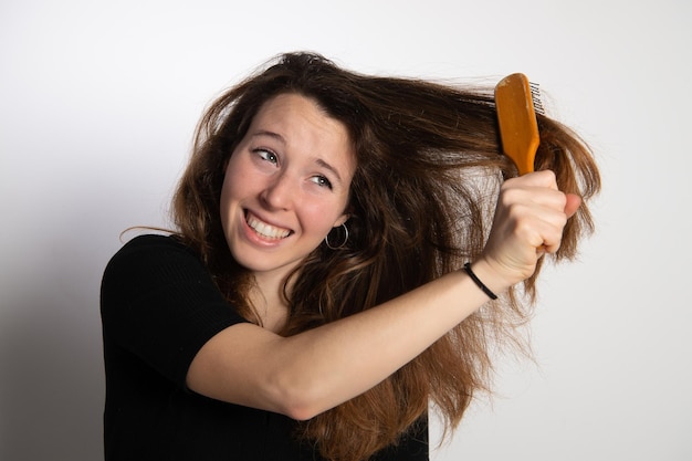 Foto bonita joven morena tratando de peinar su hermoso cabello largo y castaño con un cepillo en una mano emociones expresión dolorosa en la cara aislada en fondo blancoxdxa