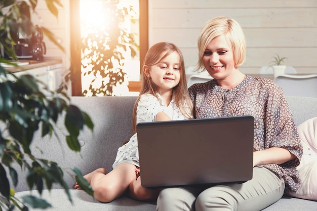 Bonita madre rubia y linda niña morena sentada en el sofá y usando la computadora portátil
