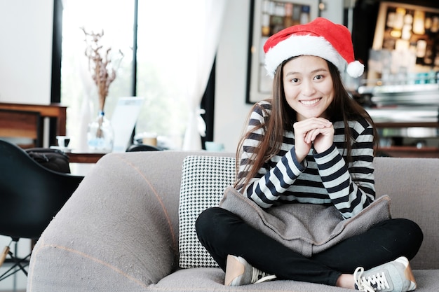 Bonita menina asiática com chapéu de natal com rosto sorridente sentado no sofá