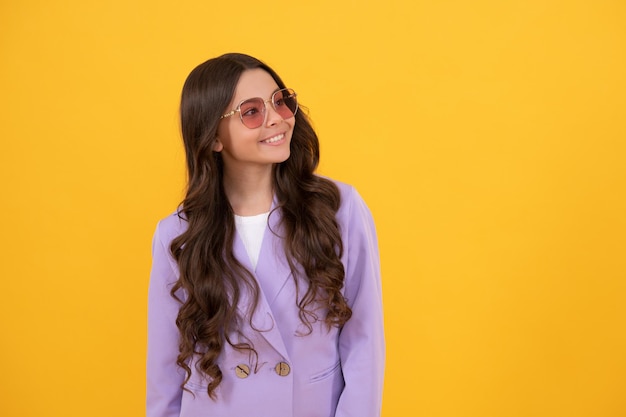 Bonita mirada de niña en chaqueta. retrato de niño feliz sobre fondo amarillo. expresar emociones positivas. niño alegre con pelo rizado en gafas de sol. belleza y moda. modelo de moda femenina.