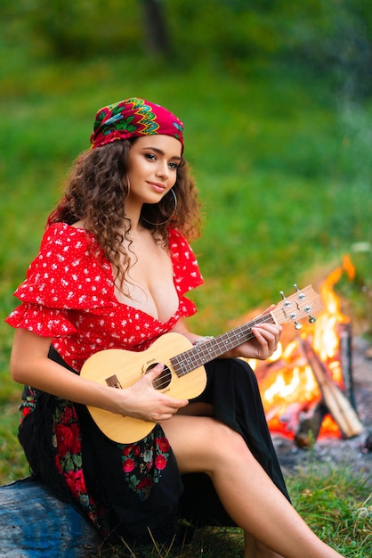 Foto bonita morena rizada con traje gitano rojo y accesorios tocando la guitarra