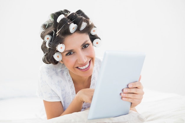 Bonita morena en los rodillos de pelo acostado en su cama con su tableta sonriendo a la cámara