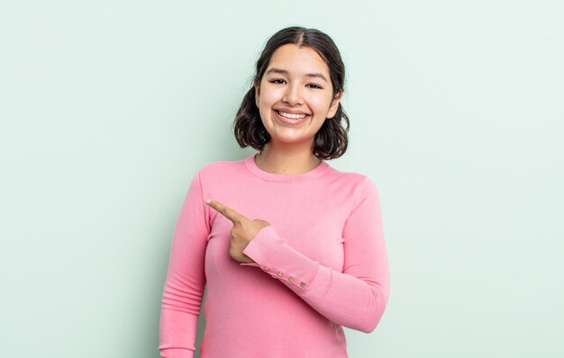 Bonita mujer adolescente sonriendo alegremente sintiéndose feliz y apuntando hacia un lado y hacia arriba mostrando objetos en el espacio de la copia