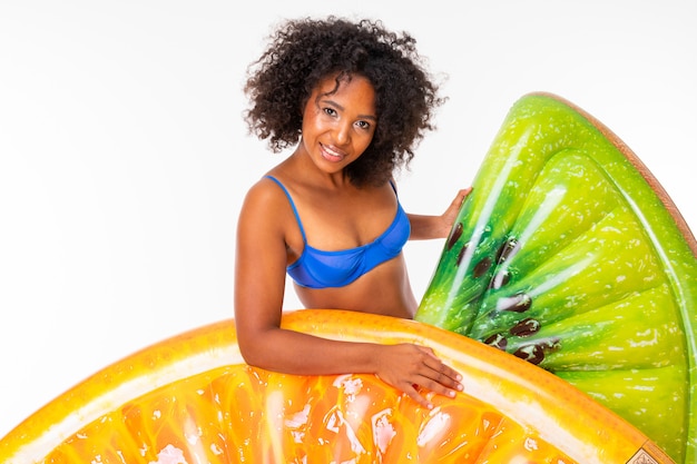 Bonita mujer africana se encuentra en traje de baño con colchones de goma playa kiwi y naranja y sonrisas aisladas