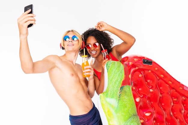 Bonita mujer africana y hombre rubio caucásico se encuentra en traje de baño con colchones de goma de playa, bebe jugo y hace selfie juntos aislado sobre fondo blanco.