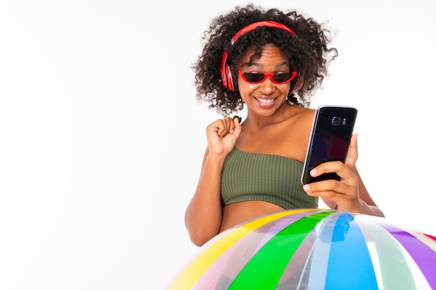 Bonita mujer africana en traje de baño se encuentra con una gran pelota de goma colorida, escucha música con auriculares y haz selfie aislado
