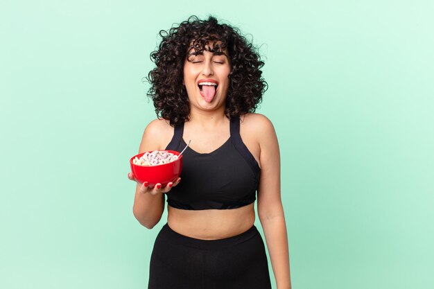 Bonita mujer árabe con actitud alegre y rebelde, bromeando y sacando la lengua vistiendo ropa de fitness y sosteniendo un tazón de copos de maíz