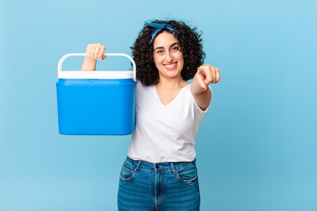 Bonita mujer árabe apuntando a la cámara eligiéndote y sosteniendo un refrigerador portátil