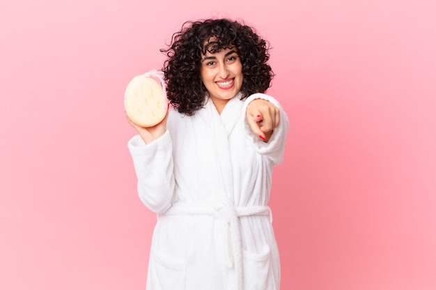 Bonita mujer árabe apuntando a la cámara eligiéndote vistiendo albornoz y sosteniendo una esponja