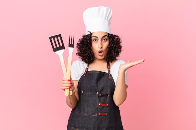 Bonita mujer árabe que parece sorprendida y consternada, con la mandíbula caída sosteniendo un objeto. concepto de chef de barbacoa