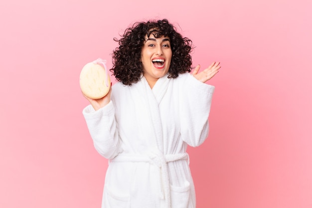 Bonita mujer árabe que se siente feliz y asombrada por algo increíble vistiendo una bata de baño y sosteniendo una esponja