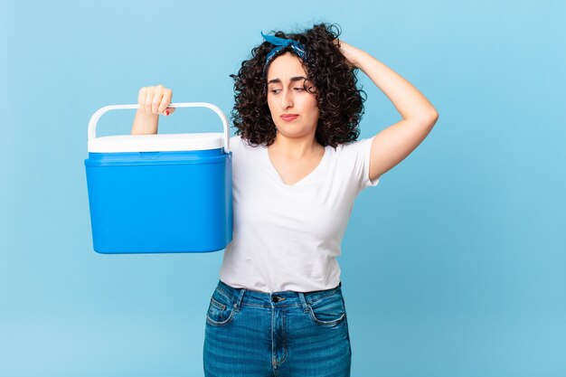 Bonita mujer árabe que se siente perpleja y confundida, rascándose la cabeza y sosteniendo un refrigerador portátil