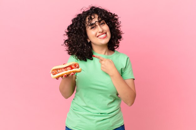 Bonita mujer árabe sonriendo alegremente, sintiéndose feliz y apuntando hacia un lado y sosteniendo un perrito caliente