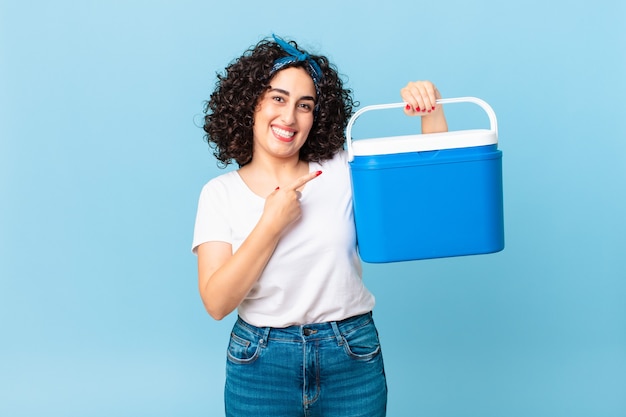 Bonita mujer árabe sonriendo alegremente, sintiéndose feliz y apuntando hacia un lado y sosteniendo un refrigerador portátil