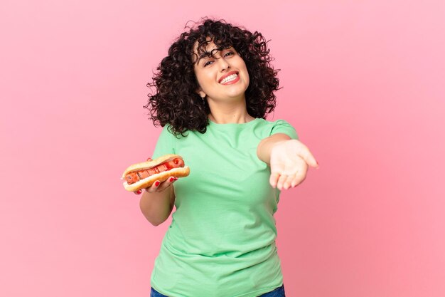 Bonita mujer árabe sonriendo felizmente con amable y ofreciendo y mostrando un concepto y sosteniendo un hot dog