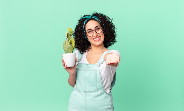 Bonita mujer árabe sonriendo felizmente con amigable y ofreciendo y mostrando un concepto y sosteniendo un cactus en maceta