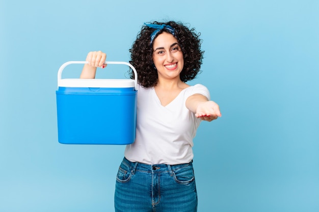 Bonita mujer árabe sonriendo felizmente con amigable y ofreciendo y mostrando un concepto y sosteniendo un refrigerador portátil