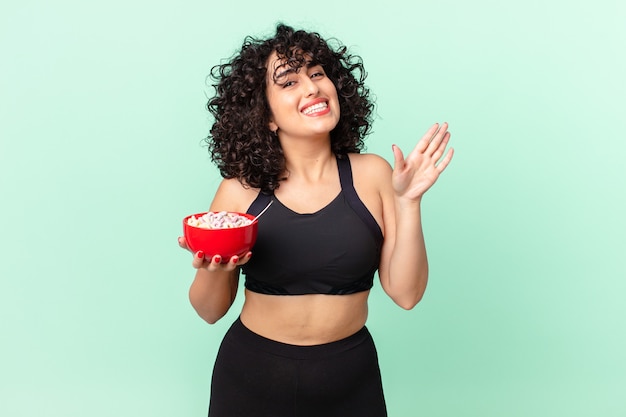 Bonita mujer árabe sonriendo felizmente, saludando con la mano, dándote la bienvenida y saludándote vistiendo ropa deportiva y sosteniendo un tazón de copos de maíz