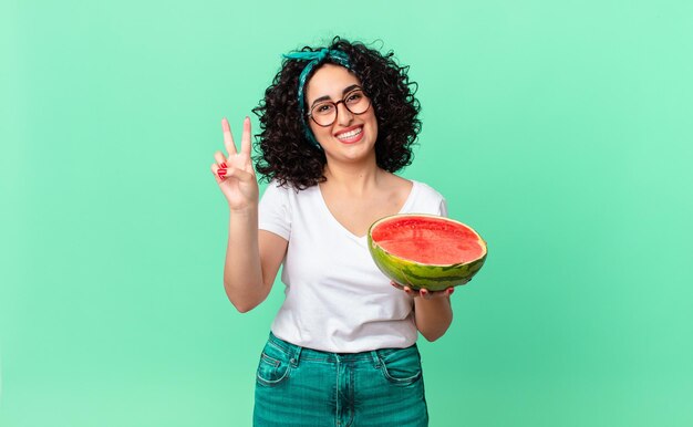 Bonita mujer árabe sonriendo y mirando amigable, mostrando el número dos y sosteniendo una sandía. concepto de verano