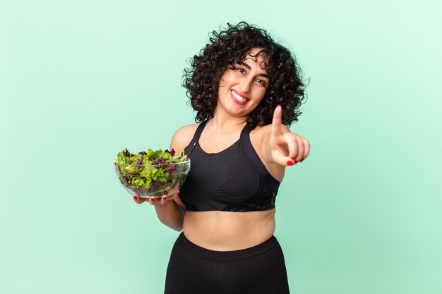 Bonita mujer árabe sonriendo con orgullo y confianza haciendo el número uno y sosteniendo una ensalada. concepto de dieta