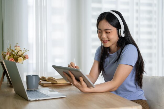 Una bonita mujer asiática escuchando música a través de sus auriculares mientras usa su tableta en una mesa