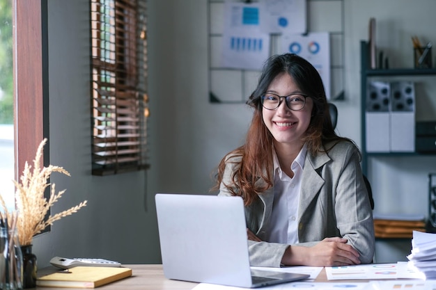 Bonita mujer asiática sentada en la calculadora de la oficina trabajando en gráficos de datos y documentos en la mesa en el espacio de trabajo de negocios mirando la cámara