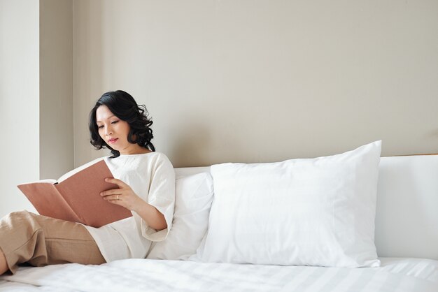Bonita mujer asiática sentada en la cama y leyendo un libro interesante